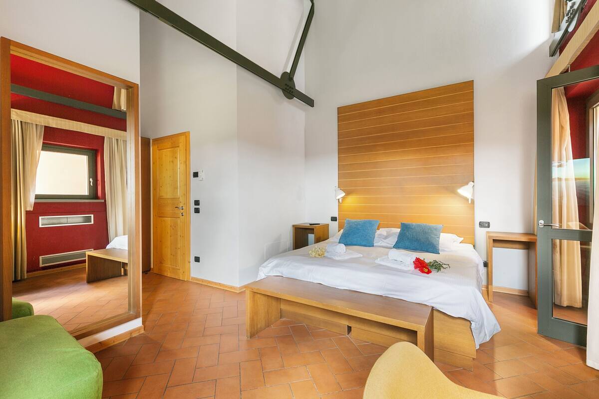 Habitación de hotel con cama, mesitas de noche, un banco, cabecera de madera y una ventana con cortinas.