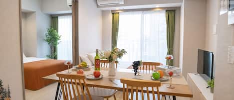 Una habitación de hotel moderna con zona de comedor, cama, televisor y ventana con cortinas.