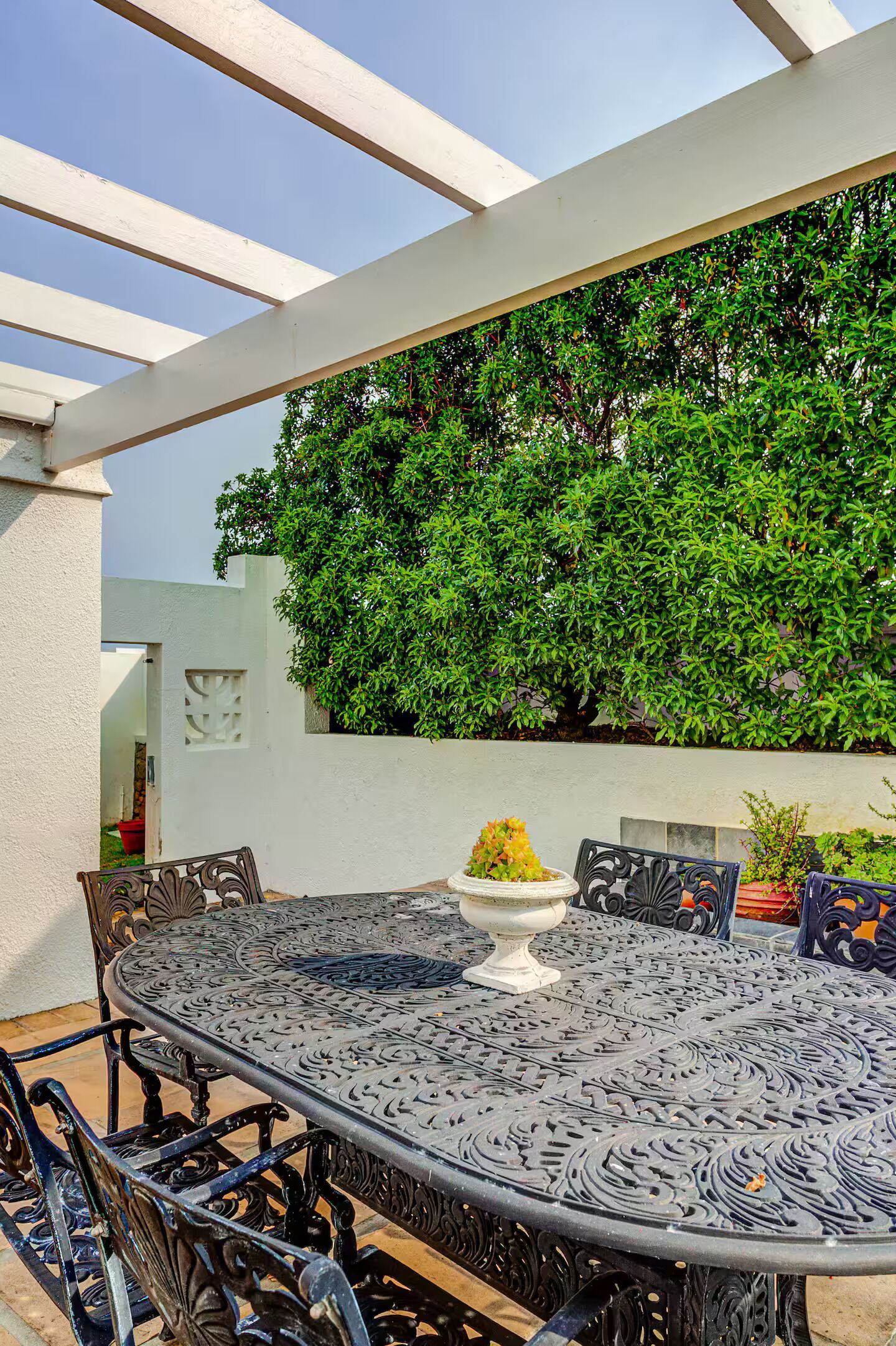 Eine Terrasse im Freien mit einem runden Metalltisch und Stühlen, umgeben von einer weißen Mauer und einem großen, grünen Busch.