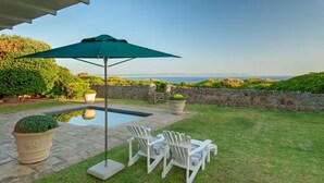 A backyard with a swimming pool, two white Adirondack chairs, a green umbrella, and potted plants. - 4-bedroom house overlooking the sea on the cliff path in Hermanus (Hermanus)