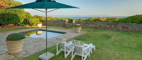 A backyard with a swimming pool, two white Adirondack chairs, a green umbrella, and potted plants.
