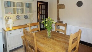 A wooden dining table with four chairs, a vase with flowers, a white cabinet with glass doors, and a stone wall.