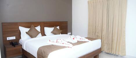 A neatly made bed with a brown headboard, white bedding, and a decorative white towel with red rose petals. There is a wooden nightstand with a phone and a power outlet. A beige curtain is partially drawn, and an air conditioning unit is mounted above the bed.