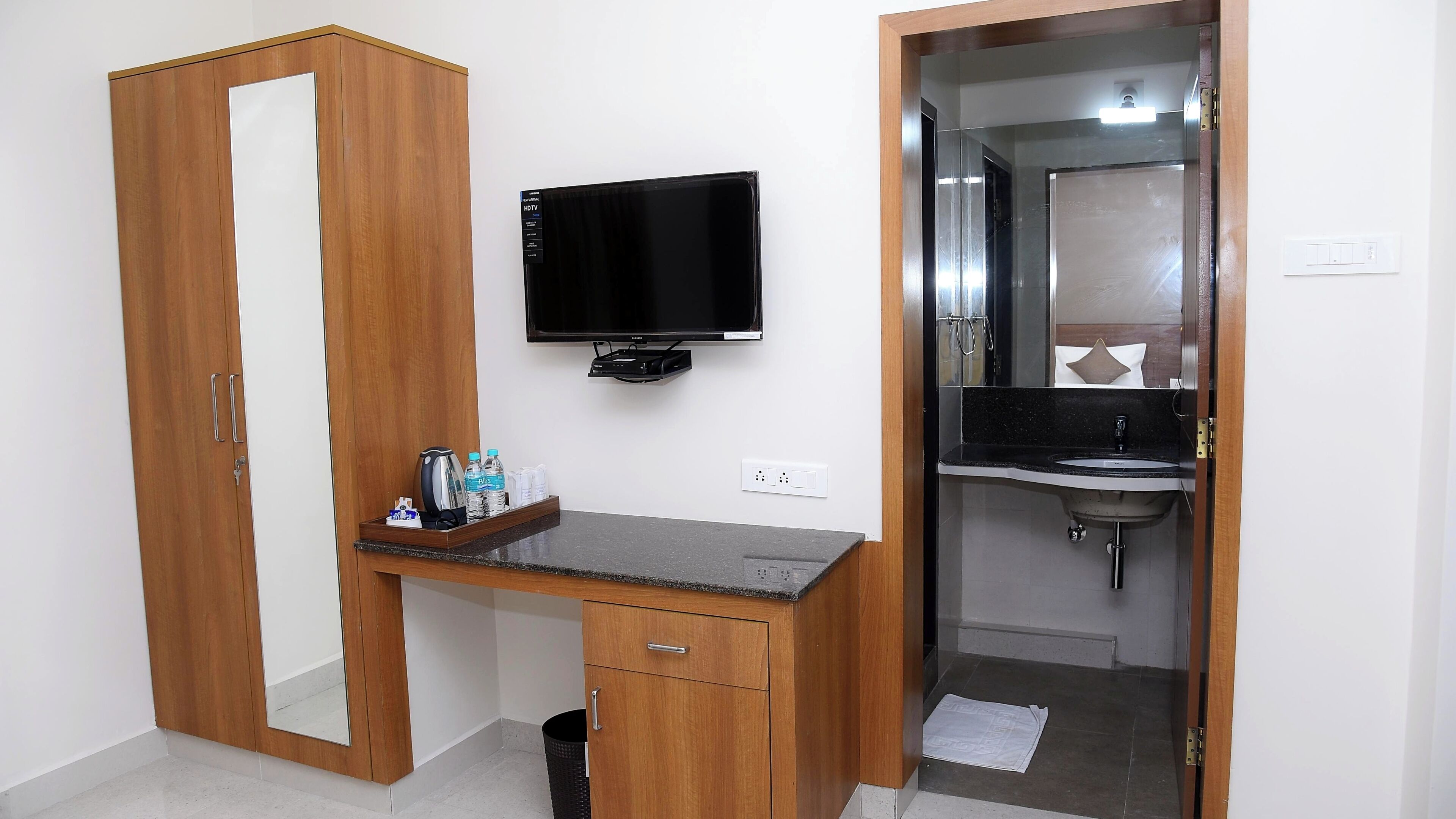 A hotel room with a wooden wardrobe, a desk with a flat-screen TV, a small shelf with toiletries, and a bathroom with a sink and mirror.