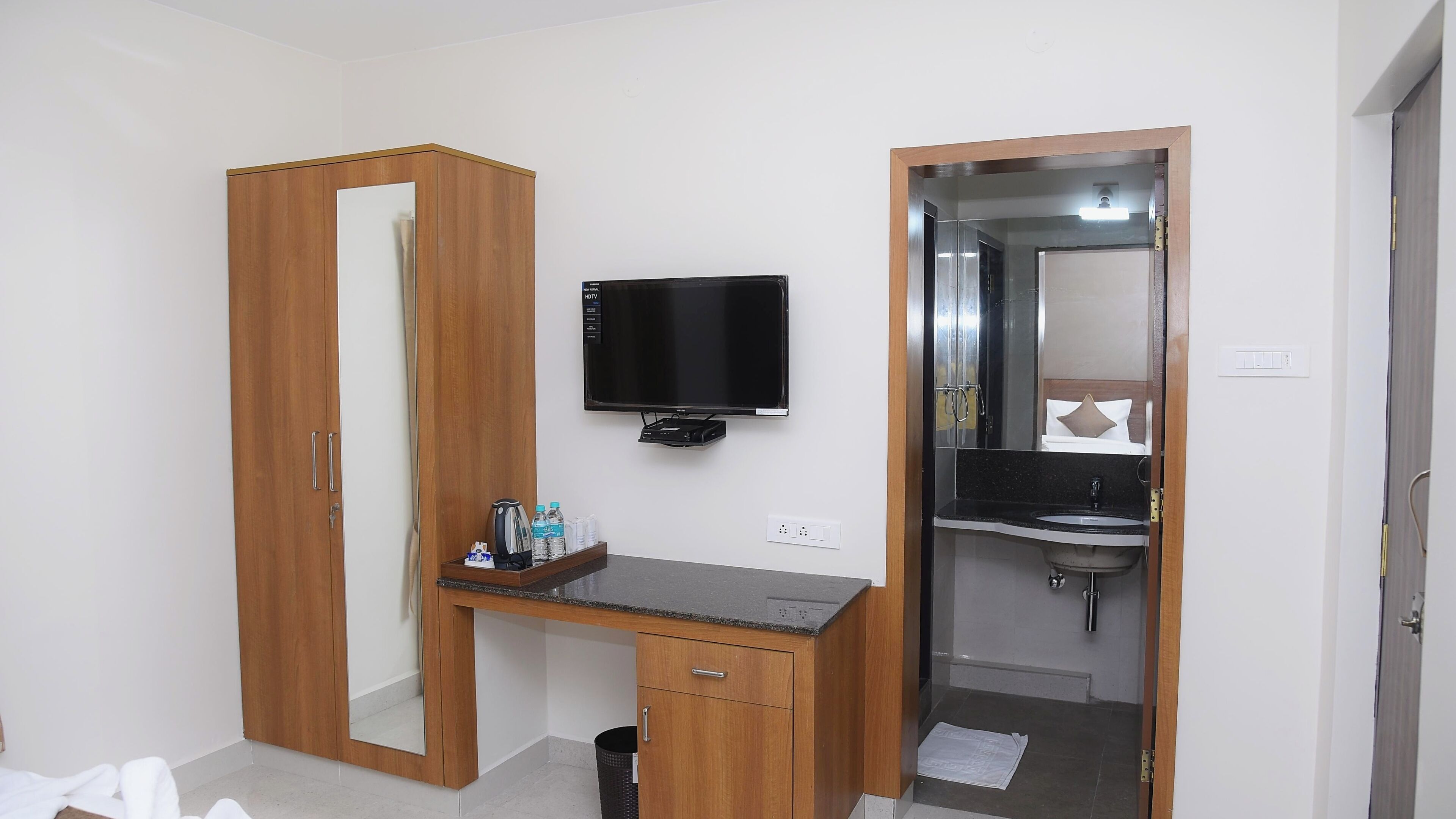 Une chambre d’hôtel avec une armoire en bois, un téléviseur fixé au mur, un petit bureau avec un miroir et une salle de bain équipée d’un lavabo et d’un miroir.