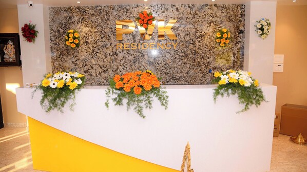 A reception desk with 'FJK RESIDENCY' signage, three flower arrangements, and a security camera above.