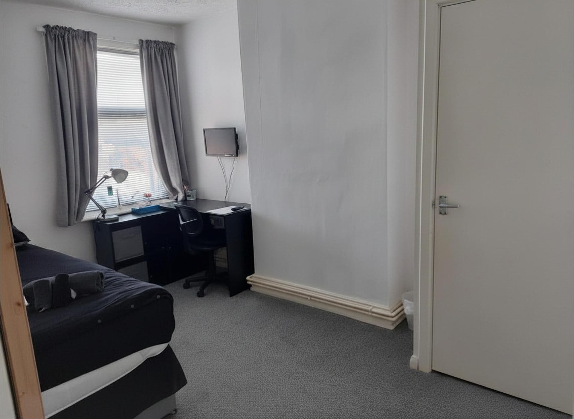 A bedroom with a bed, desk, chair, and a TV on the wall.