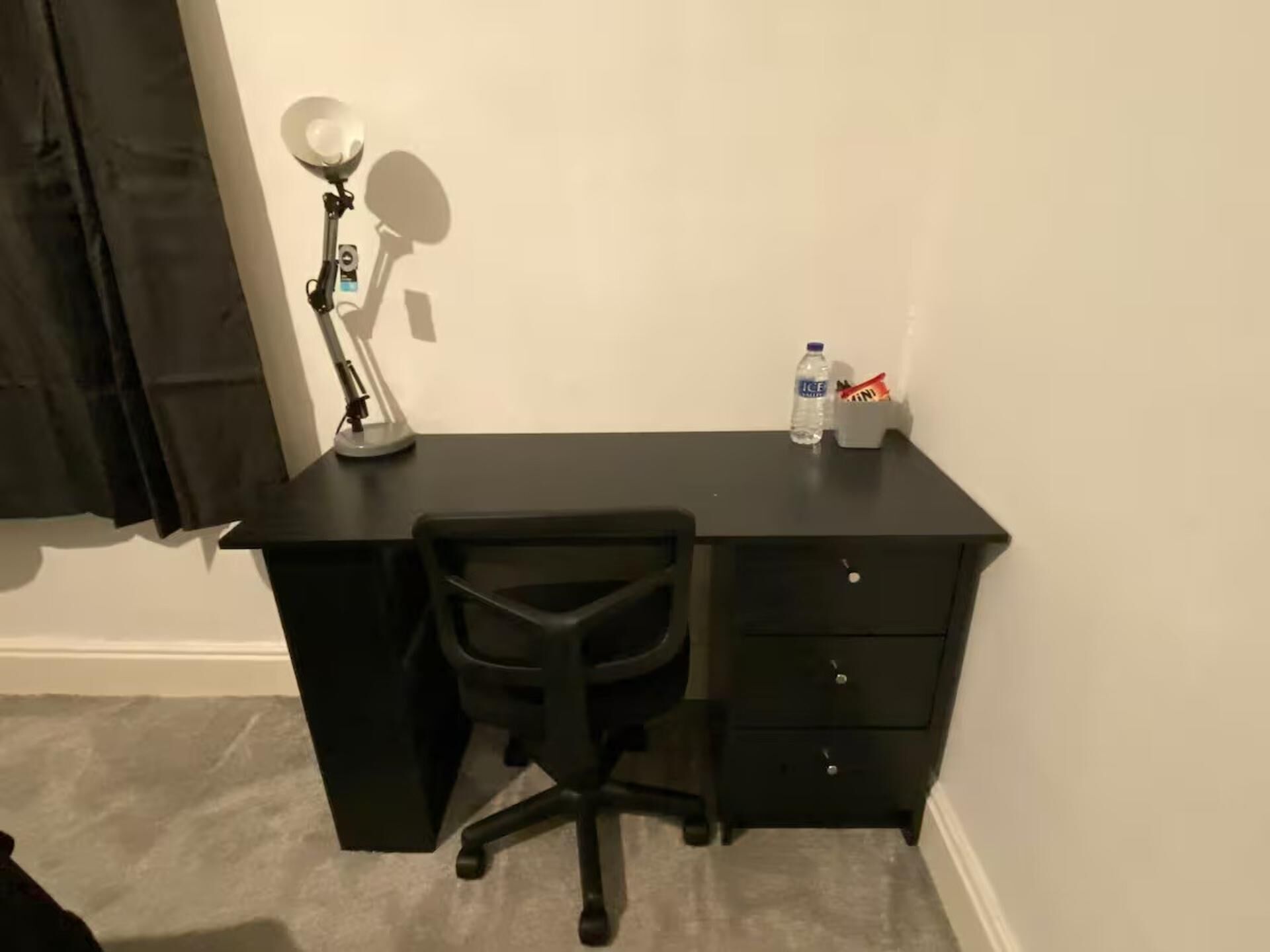 A black desk with a chair, a desk lamp, a water bottle, and a snack container.