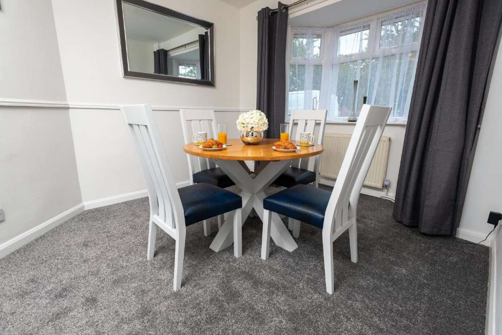 Un coin repas comprenant une table ronde, quatre chaises, un miroir et une fenêtre avec des rideaux.