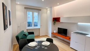 A modern, well-lit room with a dining table set for four, a green sofa, a TV, and a small kitchen area. - 4bnb - Charming Giuliana Apartment (Rome)