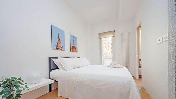 A modern bedroom with a large bed, white bedding, a bedside table with a plant, and two wall-mounted pictures. - 4bnb - Charming Giuliana Apartment (Rome)