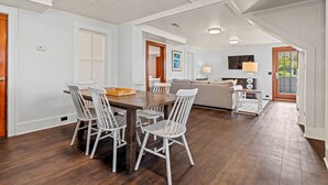 Een ruime eethoek met een houten tafel en witte stoelen, een zithoek met een bank en televisie, en een trap naar een ander niveau.