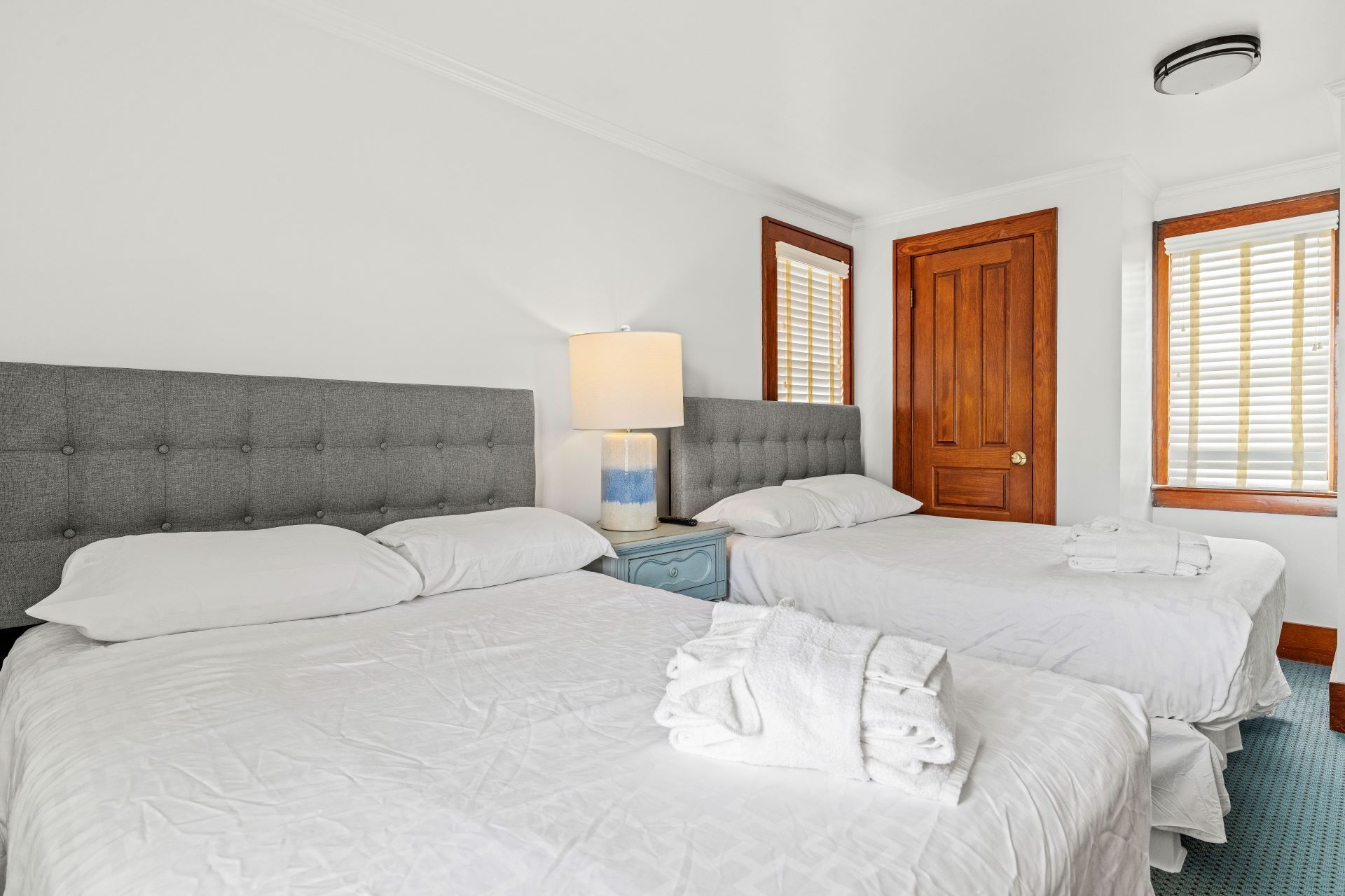 A hotel room with two beds, a grey upholstered headboard, a bedside table with a lamp, and a wooden door.