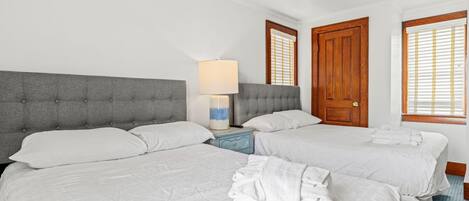 A hotel room with two beds, a grey upholstered headboard, a bedside table with a lamp, and a wooden door.