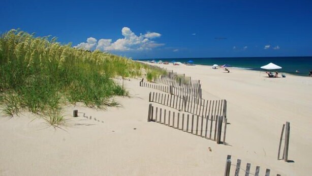 Een strand met een houten omheining, strandparasols en een helderblauwe lucht.