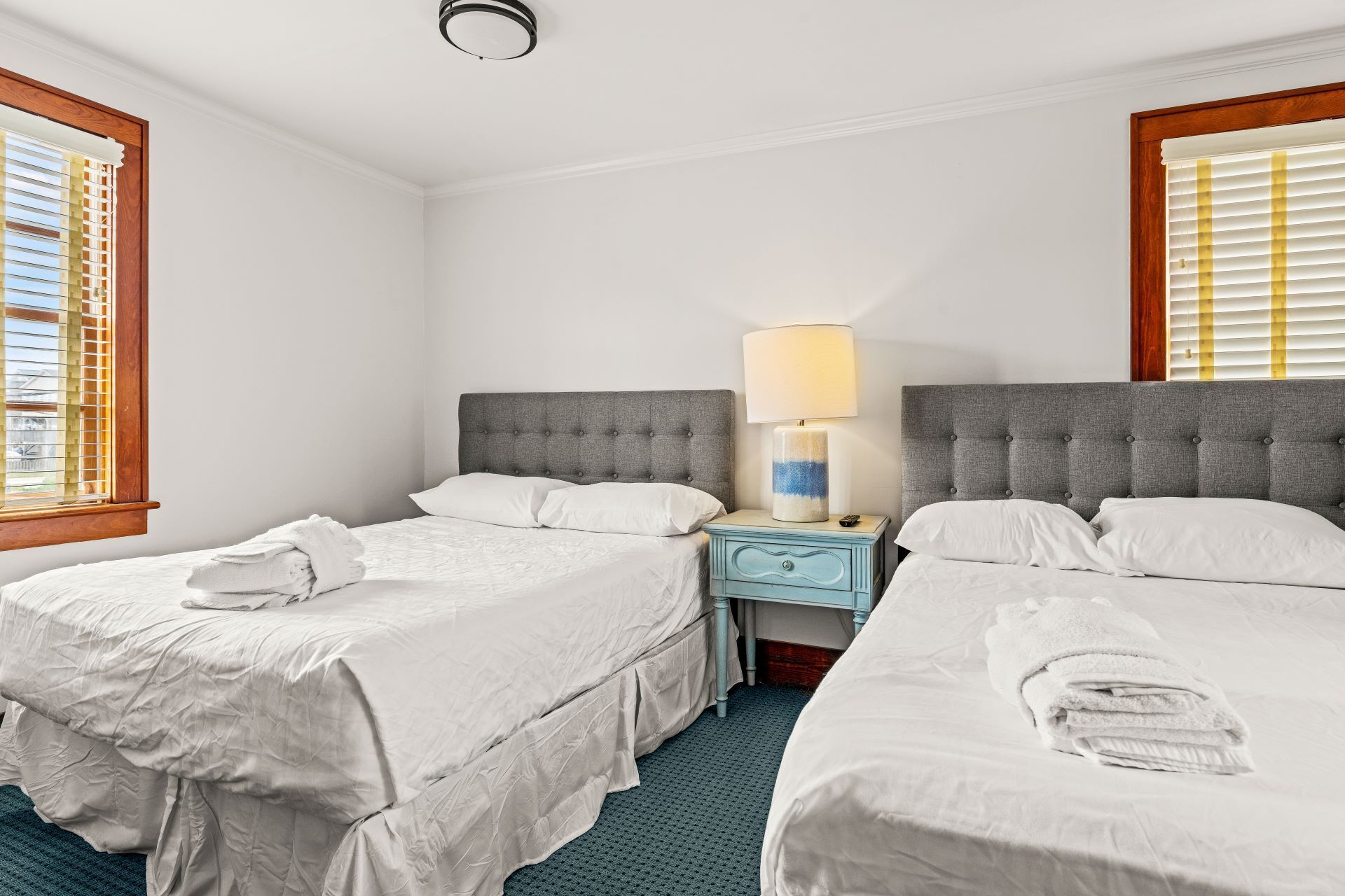 A hotel room with two beds, a nightstand with a lamp, and a window with blinds.