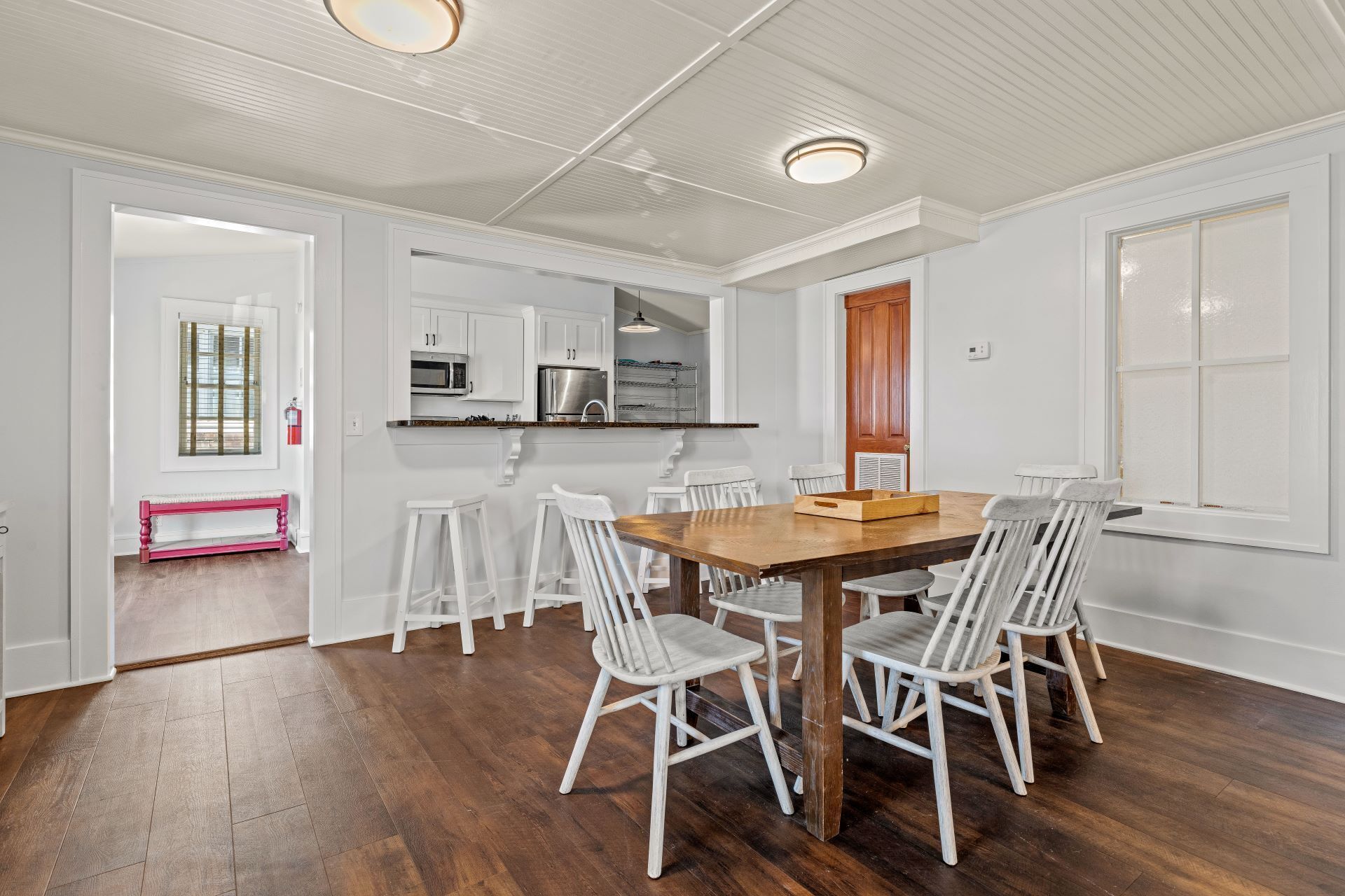 Un coin repas avec une table en bois et des chaises blanches, une cuisine avec des meubles blancs et un banc placé dans un coin.