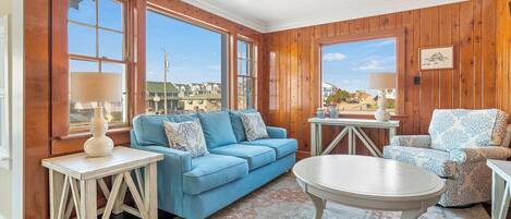 Una habitación con paneles de madera, un sofá azul, un sillón estampado, una mesa redonda y vistas a edificios a través de amplios ventanales.