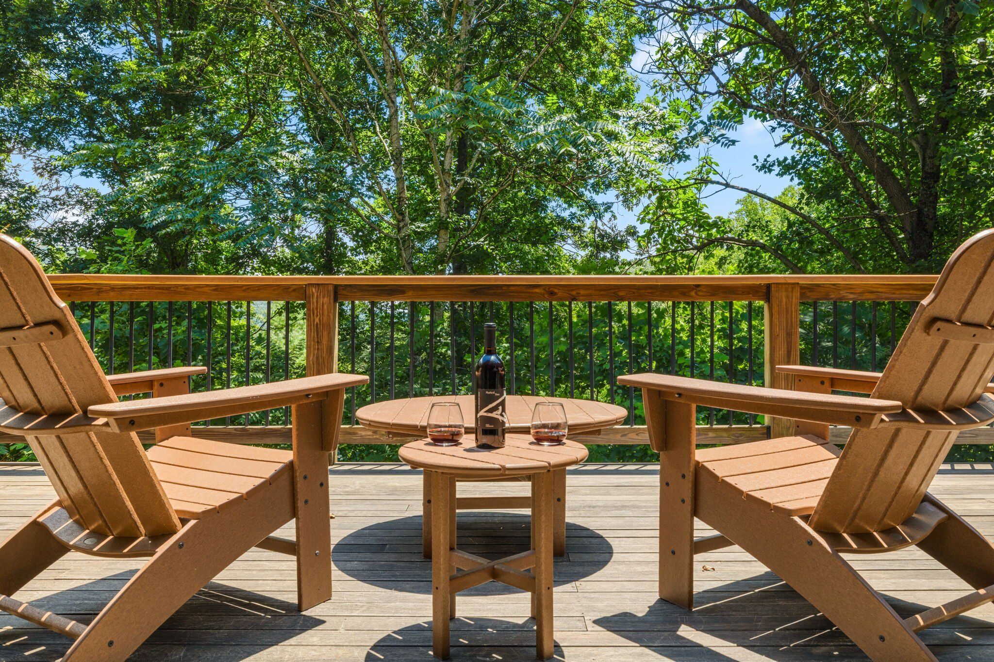 Um deck de madeira com duas cadeiras Adirondack e uma pequena mesa redonda, cada um com uma taça de vinho e uma garrafa.
