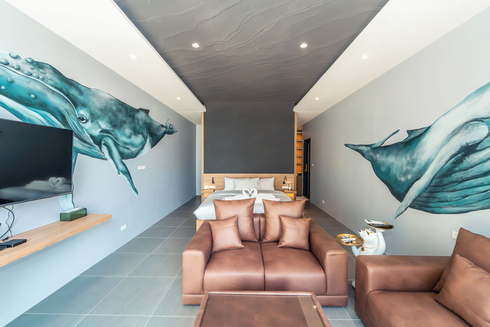 A modern hotel room with a large mural of a whale on the walls, a sofa, a TV, and a bedside table with a lamp.