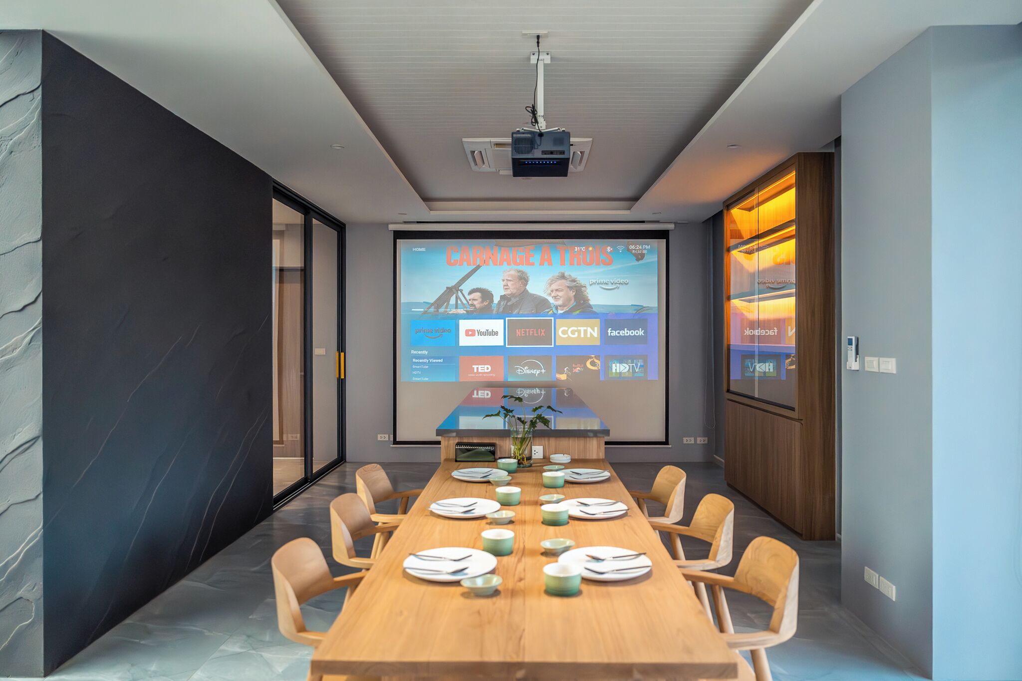 A modern conference room with a long table set for a meeting, a projector screen displaying a video, and a wooden cabinet with glass doors.