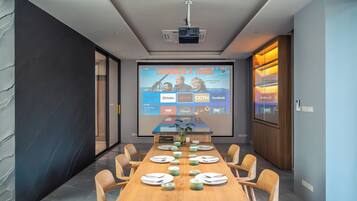 A modern conference room with a long table set for a meeting, a projector screen displaying a video, and a wooden cabinet with glass doors.