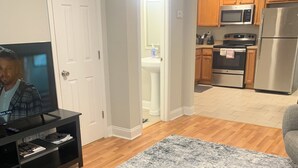 A living room with a TV, a grey rug, wooden flooring, and a kitchen with wooden cabinets and appliances. - Welcome to our Place (Charlotte)