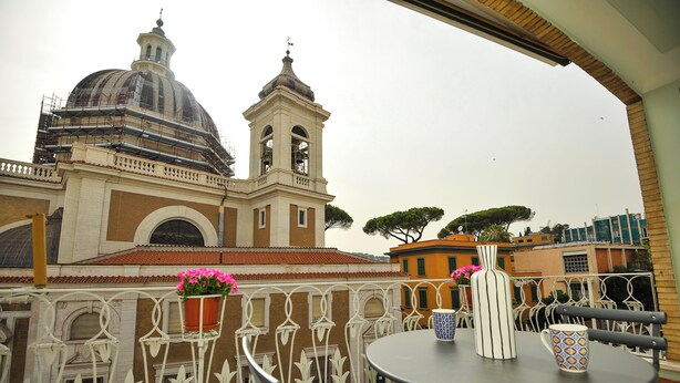 A balcony with a table and chairs, a potted plant, and a view of a historic building with a dome and bell tower. - Boutique Suite Valadier (Rome)