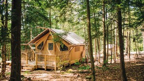 A wooden cabin with a porch and a small deck, surrounded by trees in a forest. - Huttopia Berkshires  (Williamstown)