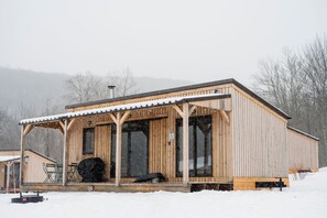 A wooden cabin with a covered porch, large windows, and a snow-covered roof. - Huttopia Berkshires  (Williamstown)