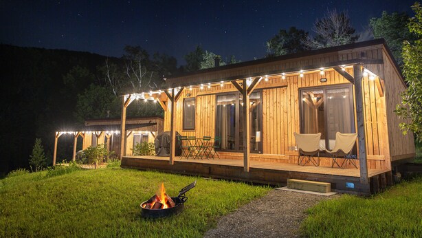A wooden cabin with a covered deck, a fire pit, and string lights. - Huttopia Berkshires  (Williamstown)