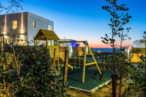 Un parque infantil con columpios y una estructura para escalar, rodeado de vegetación y edificios modernos.