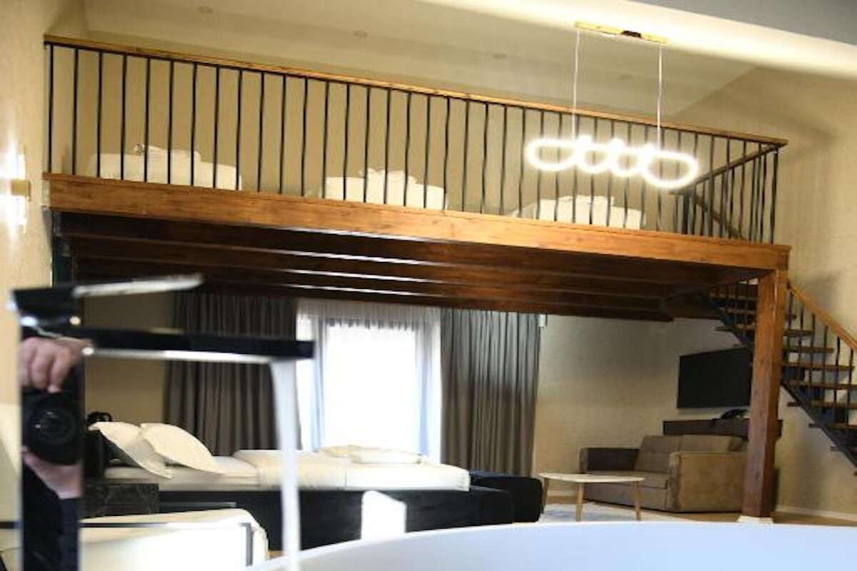 A spacious room with a large white bathtub, a wooden staircase, and a television on the wall.