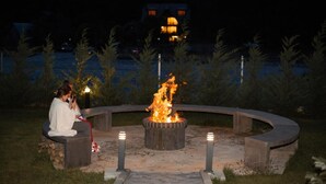 Un espacio de estar al aire libre circular con una chimenea central, rodeado por un muro de piedra bajo y pequeños árboles.