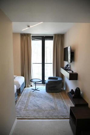 Una habitación de hotel con cama, una mesita redonda pequeña, un sofá, un televisor fijado en la pared y un ventanal con cortinas.