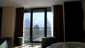 Habitación de hotel con un ventanal grande que ofrece vistas a la montaña, una cama, una mesita pequeña y dos otomanes.