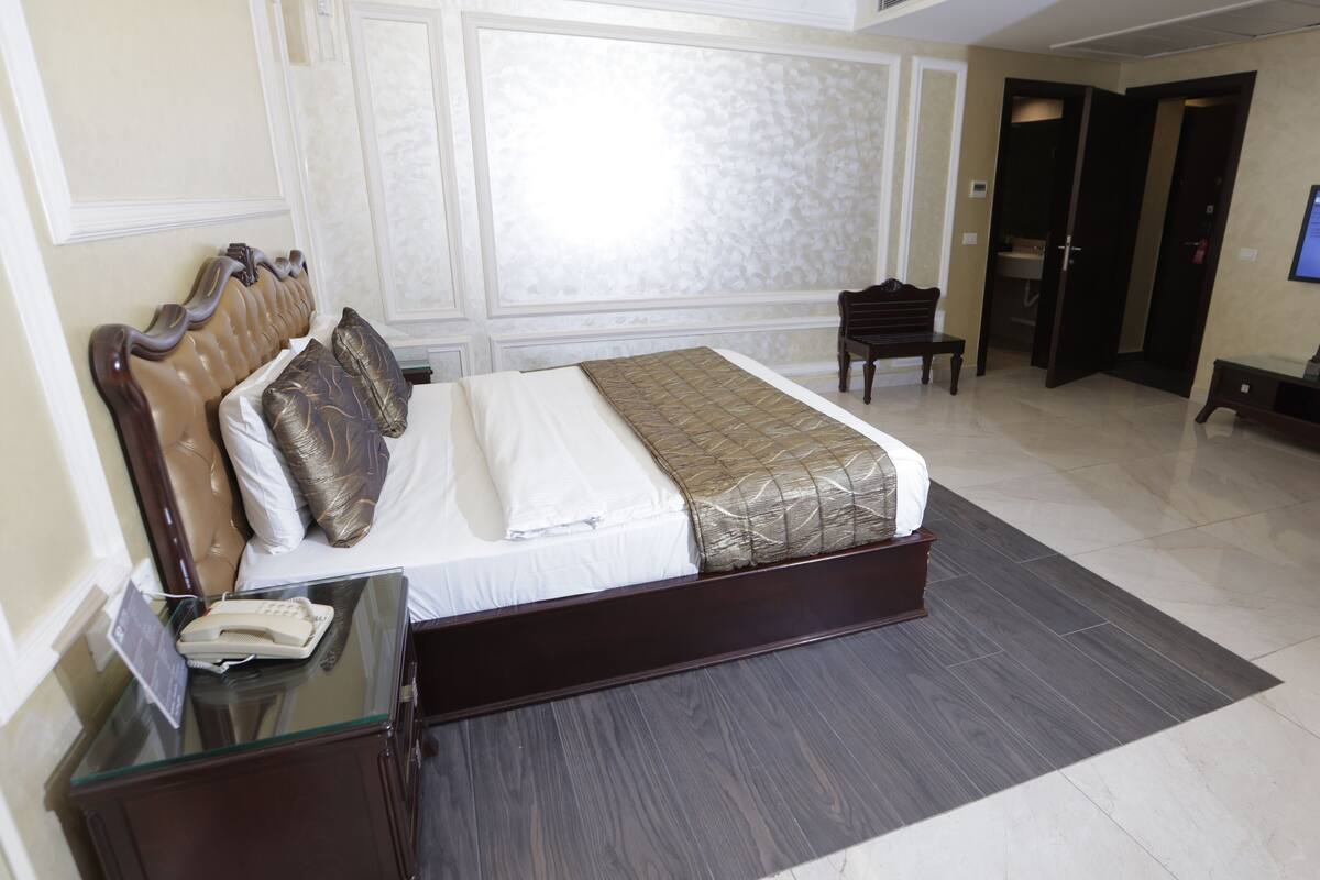 A hotel room with a large bed, a bedside table with a telephone, a chair, a desk with a computer, and a television on the wall.