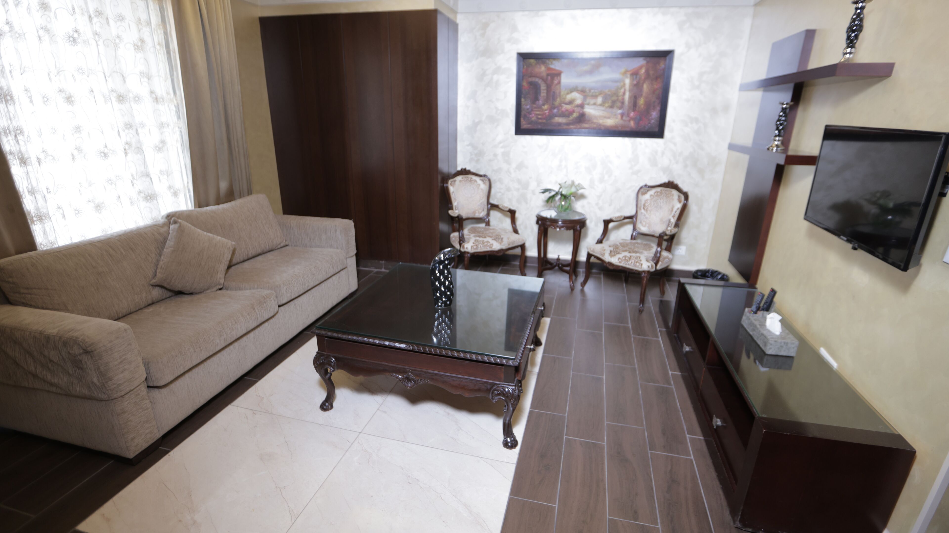 Uma sala de estar com um sofá, uma mesa de centro de vidro, duas poltronas, um quadro na parede e uma TV fixada na parede.