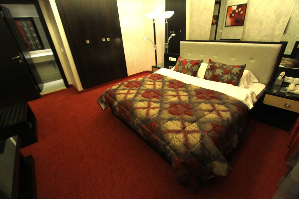 A hotel room with a large bed, a bedside table with a lamp, a nightstand, and a desk with a chair.