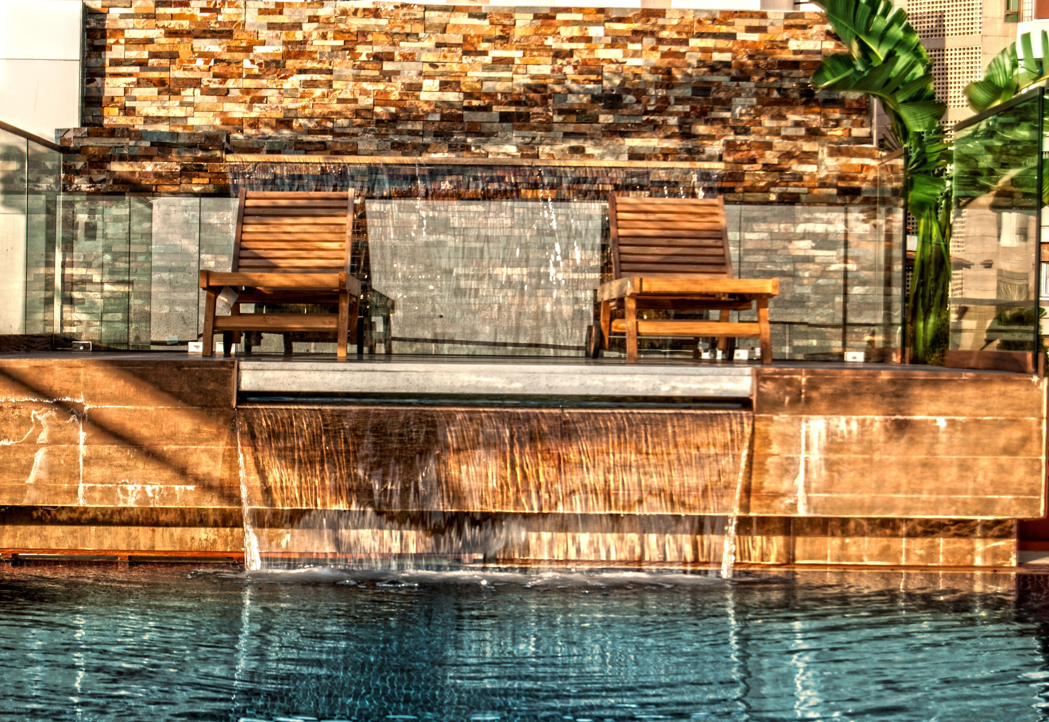 A pool area with two wooden chairs, a stone wall, and a waterfall feature.