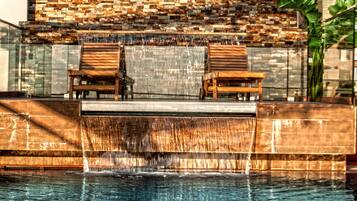 A pool area with two wooden chairs, a stone wall, and a waterfall feature.