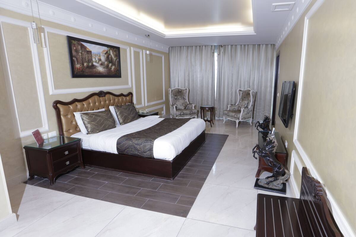A hotel bedroom with a large bed, two armchairs, a small table, a TV, and a painting on the wall.