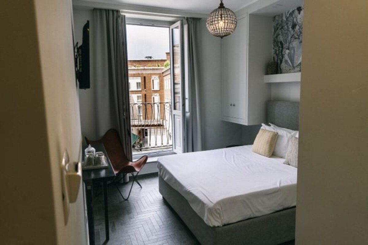 A modern hotel room with a large bed, a dining area with a chair, and a view of the city through the window.