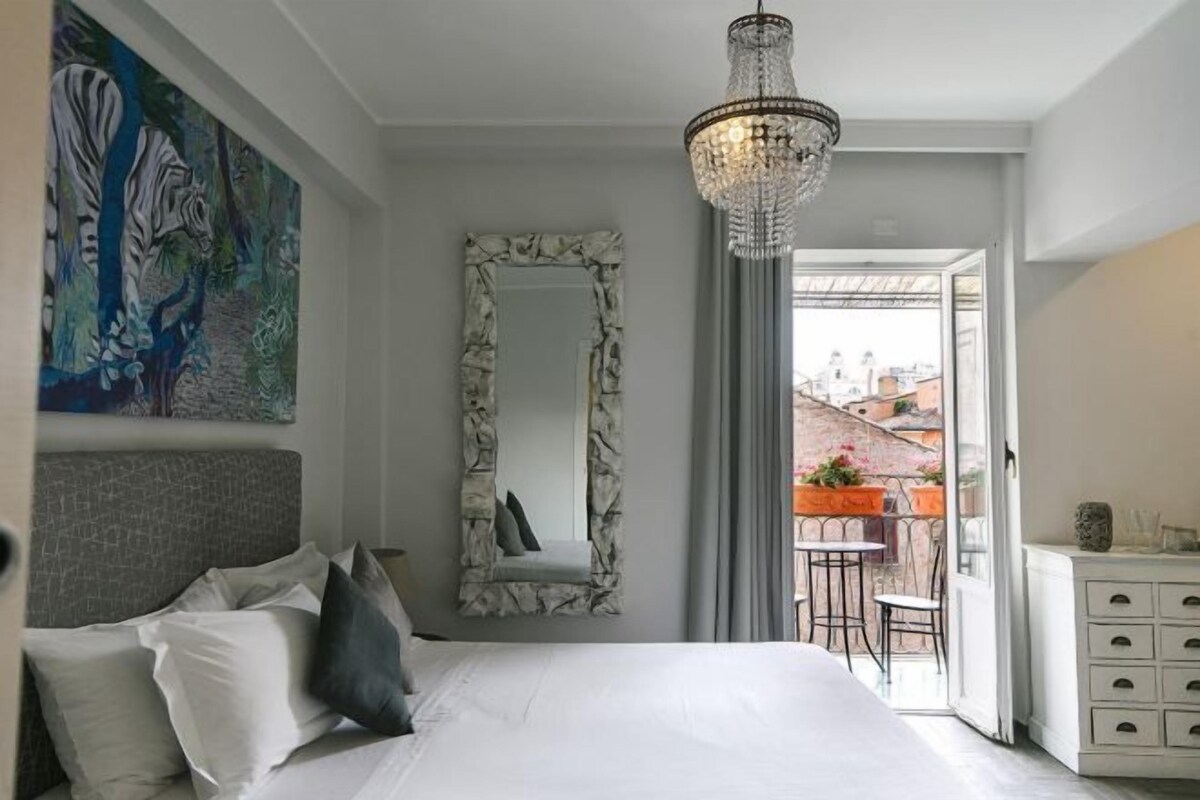 A bedroom with a large bed, a chandelier, a mirror, and a balcony with outdoor furniture.