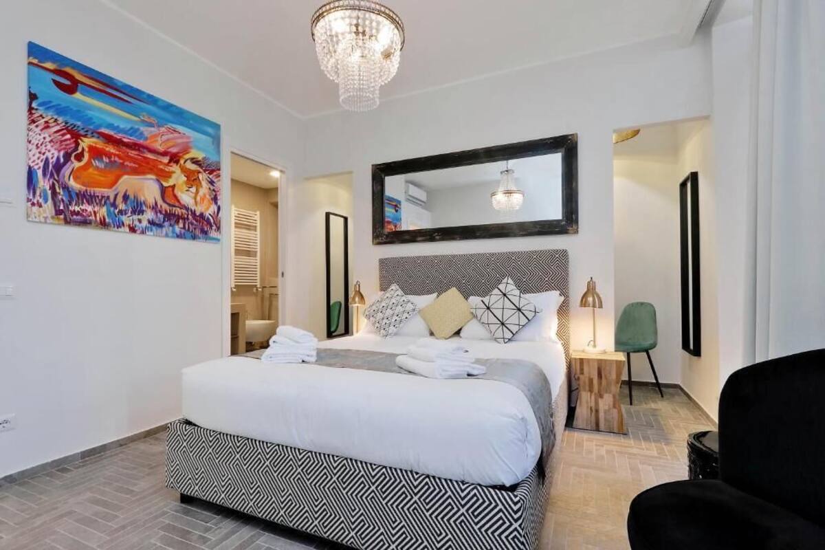 A modern hotel room with a large bed, a chandelier, a mirror, and a painting on the wall.