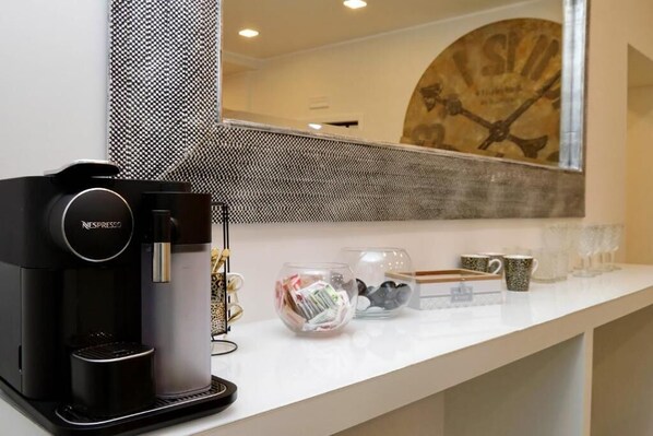 A coffee machine, a glass bowl with snacks, and a coffee maker on a white counter. - Boutique Suites della Croce (Rome)