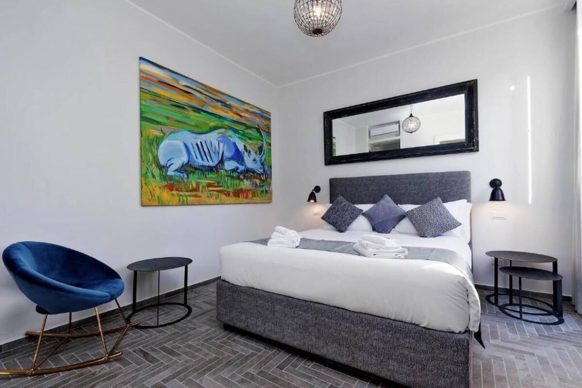 A modern bedroom with a large bed, a blue chair, a small table, a painting, and a mirror.