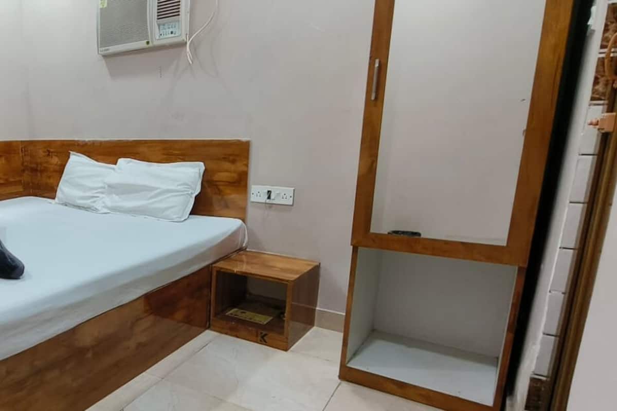 A single bed with a wooden headboard, a white mattress, and a dark blue pillow. There is a small wooden nightstand with a lamp, a power outlet, and a wall-mounted air conditioning unit. A brown curtain is partially drawn, and a door is visible to the right.