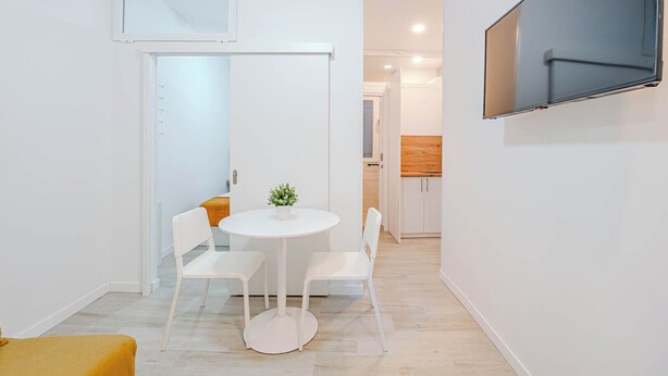 Un espacio habitable compacto con comedor, televisor montado en la pared y cocina al fondo.