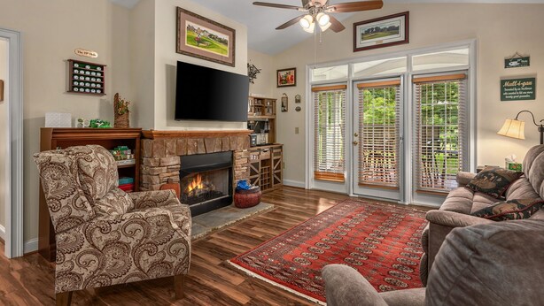 A living room with a fireplace, a television mounted above it, and a large window with blinds. - 19th Hole 3 Bedroom Condo on the 9th Green (Maggie Valley)